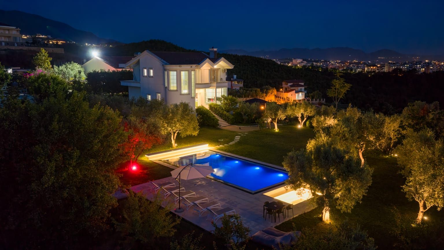 Una casa moderna con piscina, rodeada de árboles y vistas a un paisaje urbano nocturno.