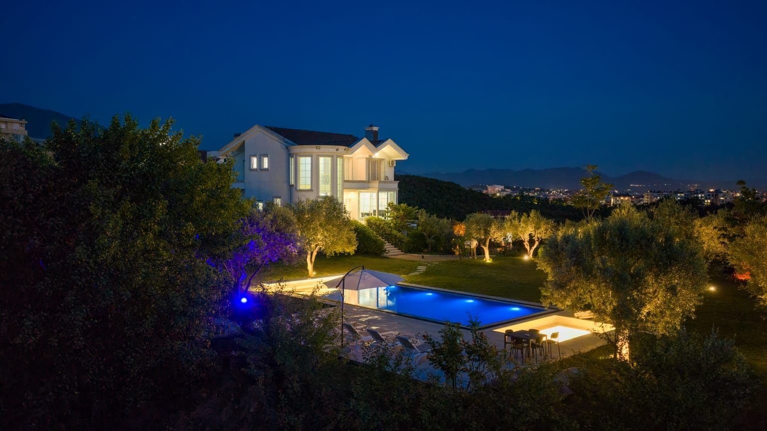 Una casa moderna con piscina, rodeada de árboles y vistas a un paisaje urbano nocturno.