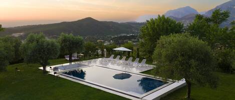 A rectangular infinity pool with white lounge chairs and a table, surrounded by greenery and trees, with a mountainous backdrop.