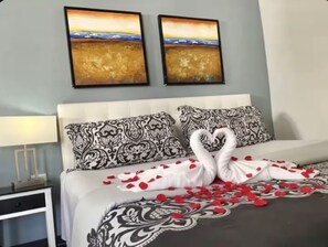 A hotel room with a bed adorned with a heart-shaped towel arrangement and rose petals, a white headboard, patterned pillows, and two framed landscape paintings on the wall. - Romantic King Studio w/ Private Jacuzzi Palm beach (Noord)