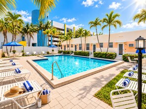 A swimming pool with lounge chairs and umbrellas, surrounded by palm trees and a building with a blue and yellow facade. - Beachside Studio I Pool I Full Kitchen (Hollywood)