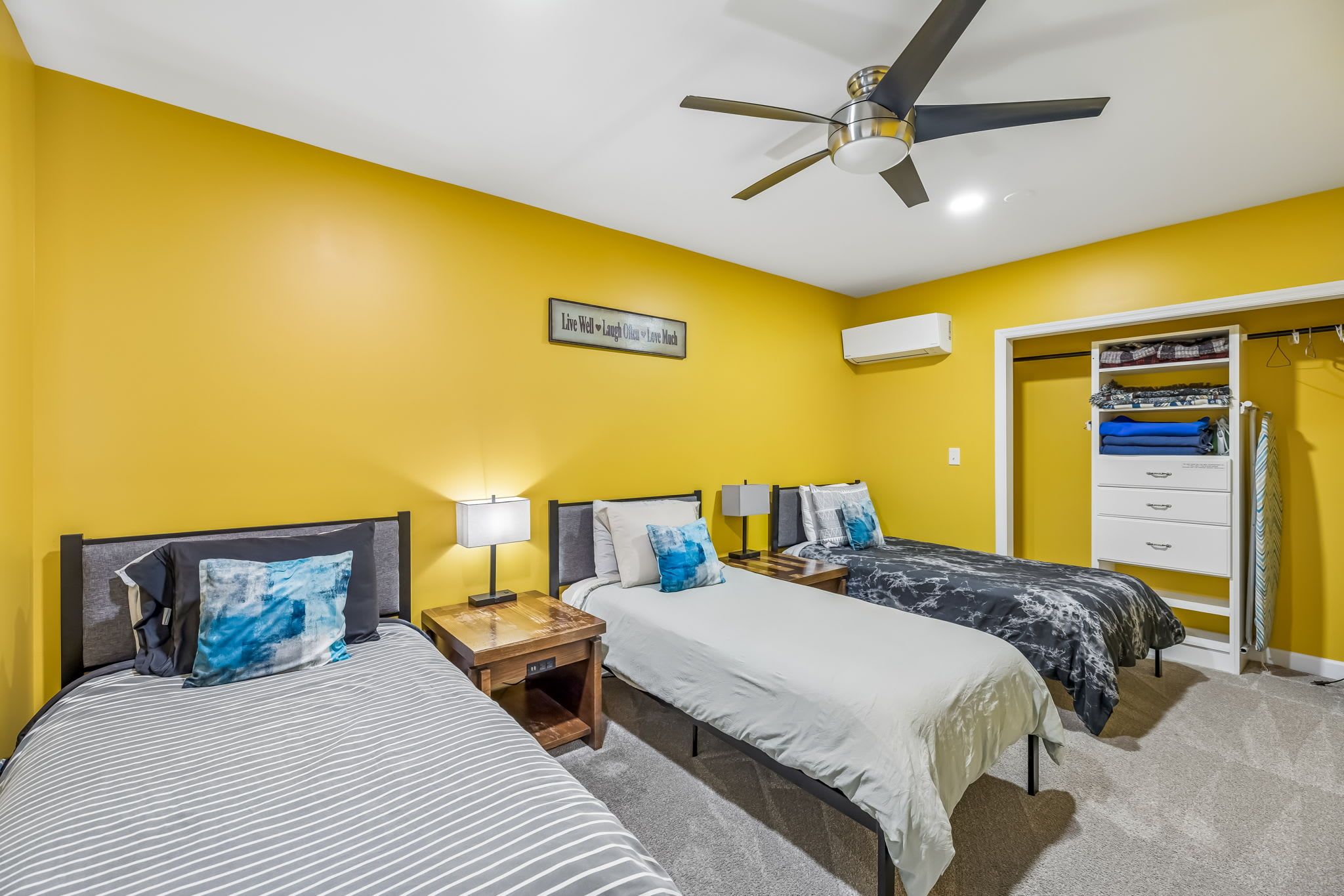 Una habitación con dos camas, ventilador de techo, aire acondicionado instalado en la pared y armario con espacio para colgar ropa.