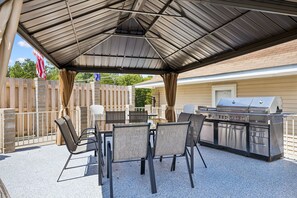 Un patio al aire libre con una zona de parrilla cubierta, una mesa con sillas y una valla de madera.