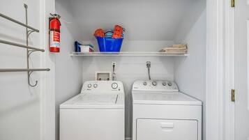 A laundry room with two white washing machines, a fire extinguisher, and a blue bucket labeled 'Travel Essentials' on a shelf.
