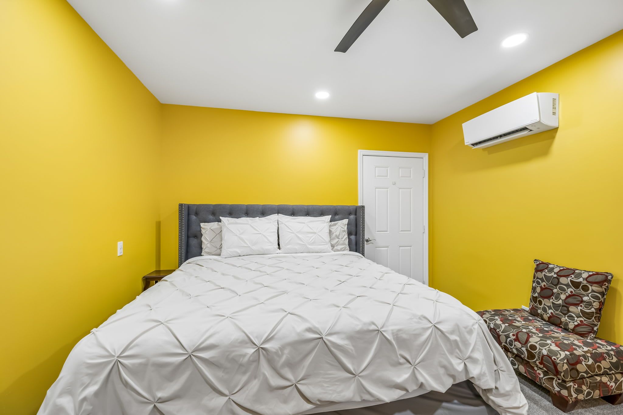 A bedroom with a bed, a chair, a door, and an air conditioning unit.