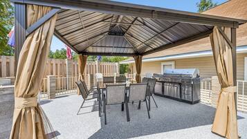 An outdoor patio with a covered grill area, seating, and a view of a house.