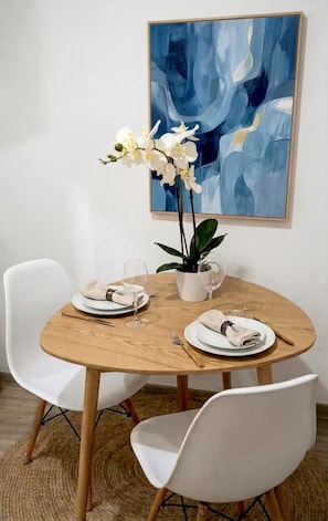 A dining area with a round wooden table, white chairs, a vase with white flowers, and a blue abstract painting on the wall. - Private 1BR Retreat • 3 Min to Palm Beach (Noord)
