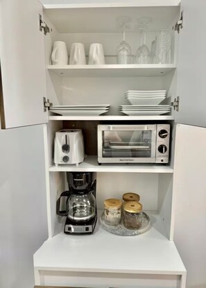 A white storage cabinet with shelves containing a toaster, a coffee maker, and jars. - Private 1BR Retreat • 3 Min to Palm Beach (Noord)