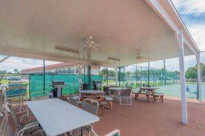 An outdoor patio area with tables and chairs, a covered section, and a view of a tennis court. - Sand, Surf & Fun / Across the street from Beach + Pool & Boat Parking (St. Augustine)