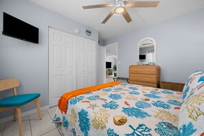 Un dormitorio con una cama, una silla, un aparador, un ventilador de techo y un televisor montado en la pared.