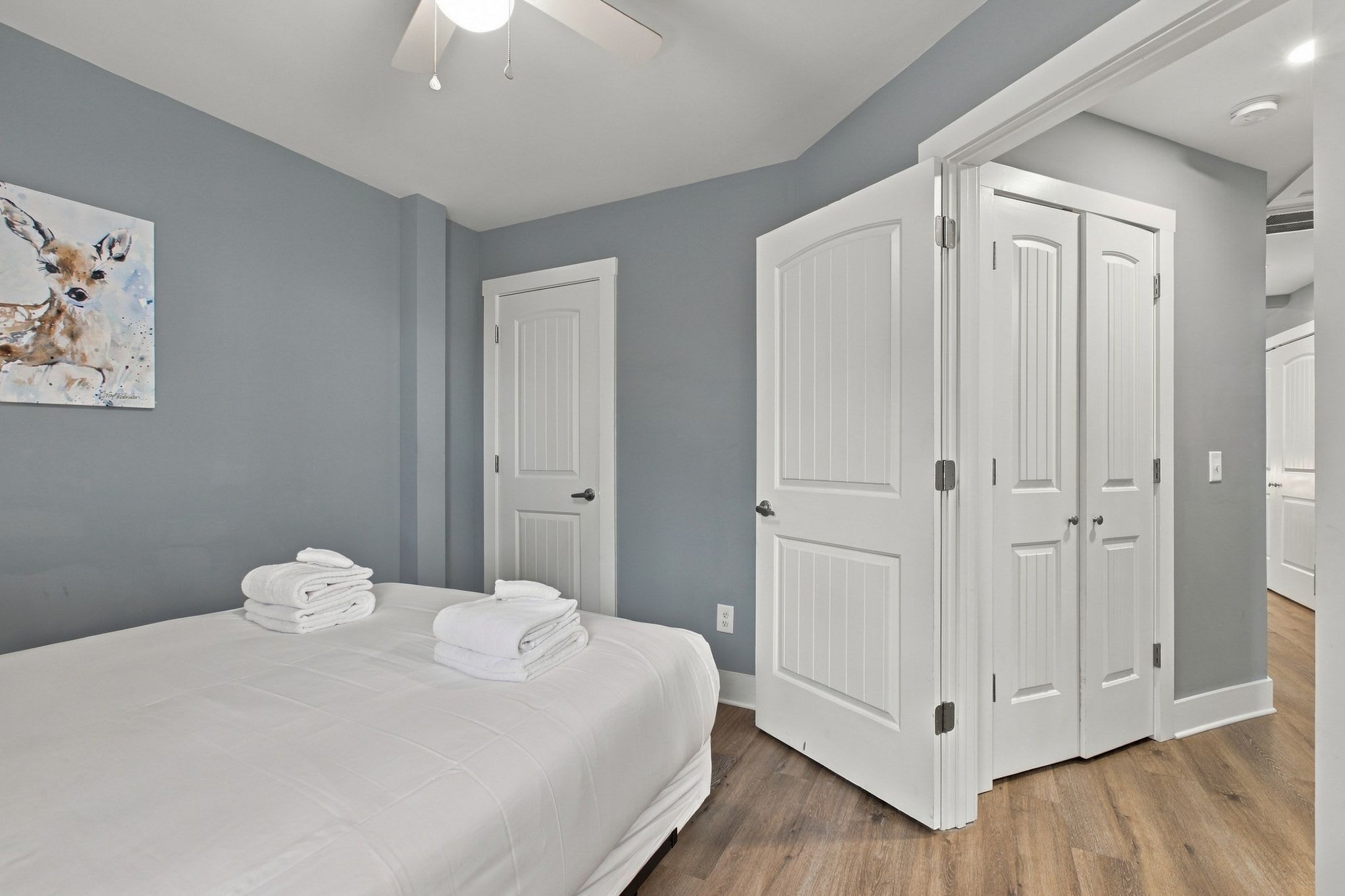 A bedroom with a bed, two doors, a ceiling fan, and a painting on the wall.