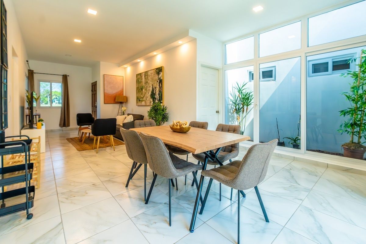 Un comedor con una mesa y sillas de madera, un cuadro en la pared y un ventanal con plantas.