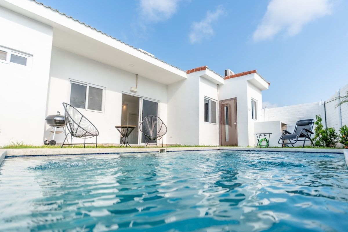 Une piscine avec une maison blanche en arrière-plan, une porte et deux chaises.