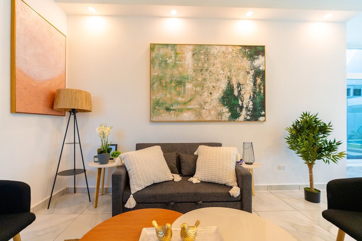 Uma sala de estar moderna com um sofá cinza, uma mesa de centro, um abajur e uma grande pintura abstrata na parede.