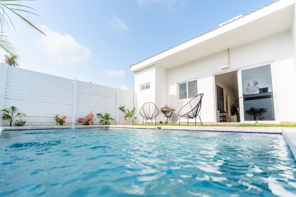 A swimming pool with a white house in the background, a covered patio area, and a white fence.