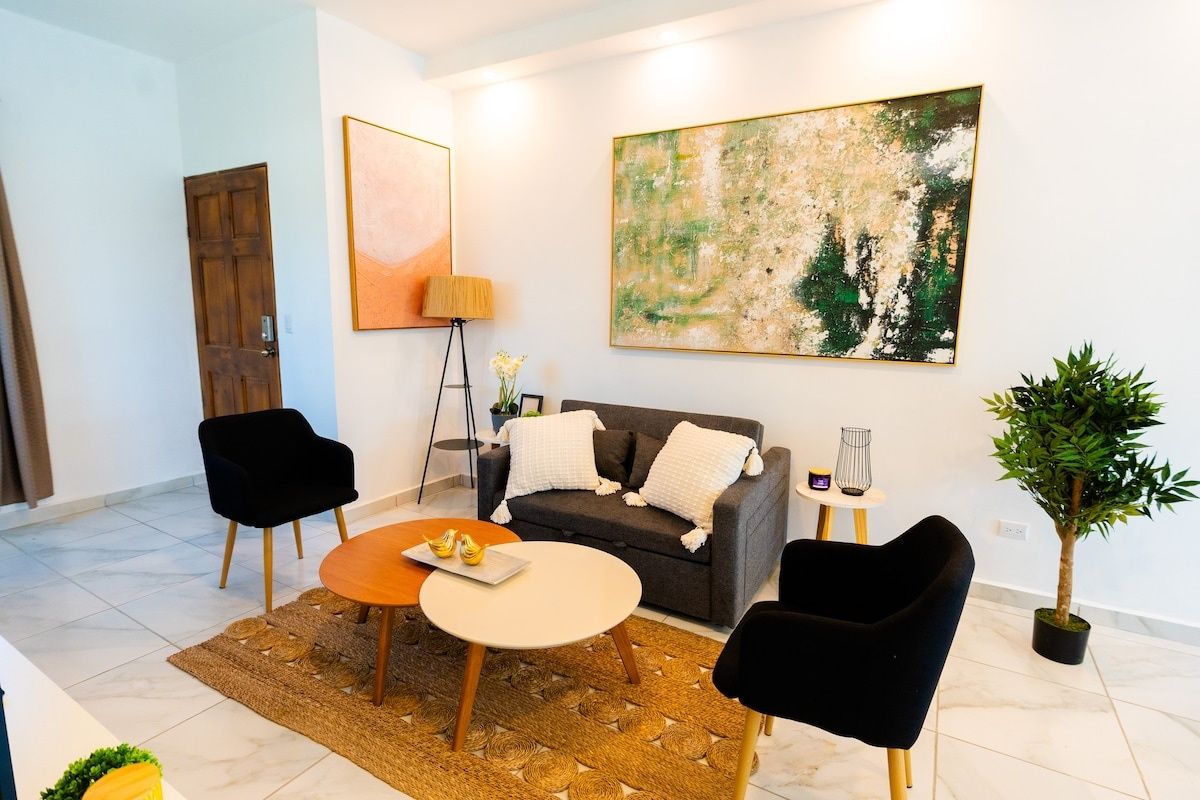 Olohuone, jossa on sohva, kaksi nojatuolia, sohvapöytä, lamppu, taulu ja ruukussa oleva kasvi.