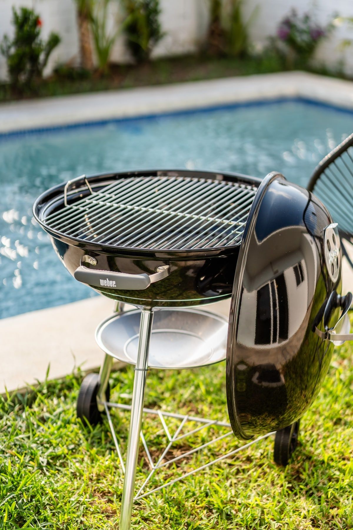 A portable Weber grill with a glass lid, placed on grass near a swimming pool.
