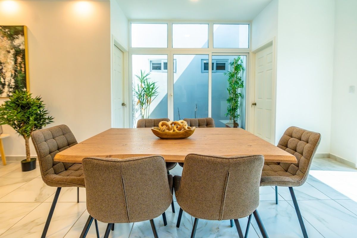 A dining area with a wooden table and four upholstered chairs, a bowl of breadsticks, and potted plants.
