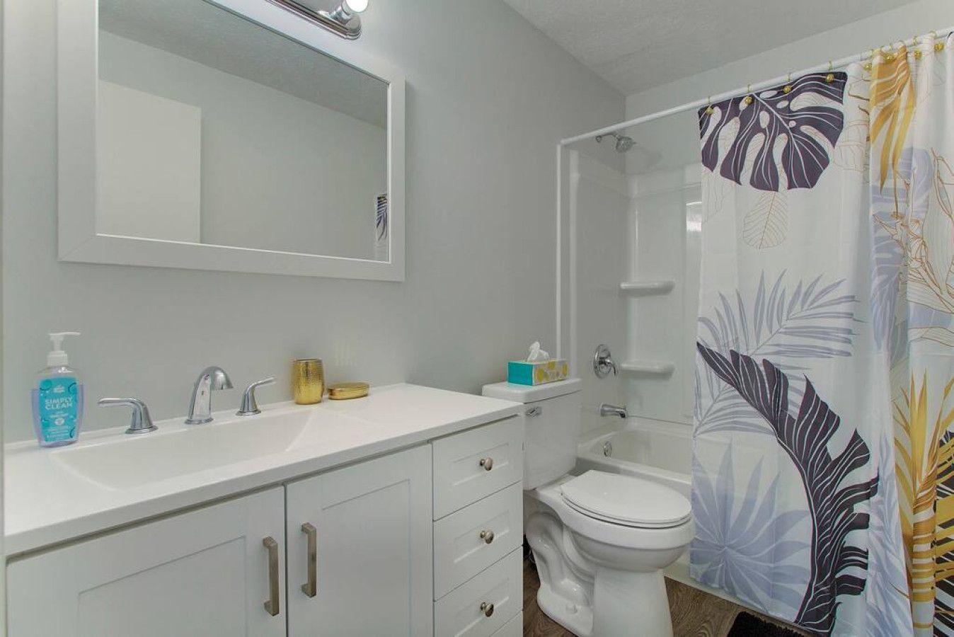 A bathroom with a white sink, two faucets, a mirror, a toilet, and a shower with a floral curtain.