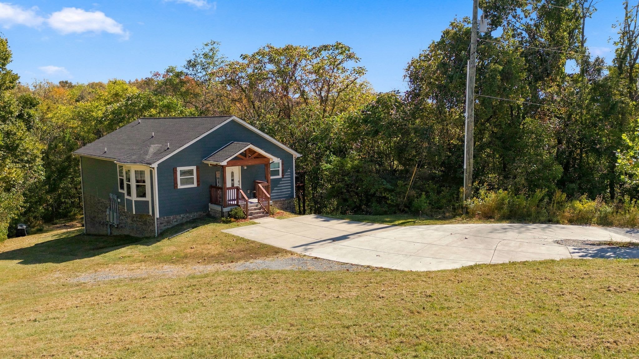 A small house with a porch, surrounded by trees and a concrete driveway.