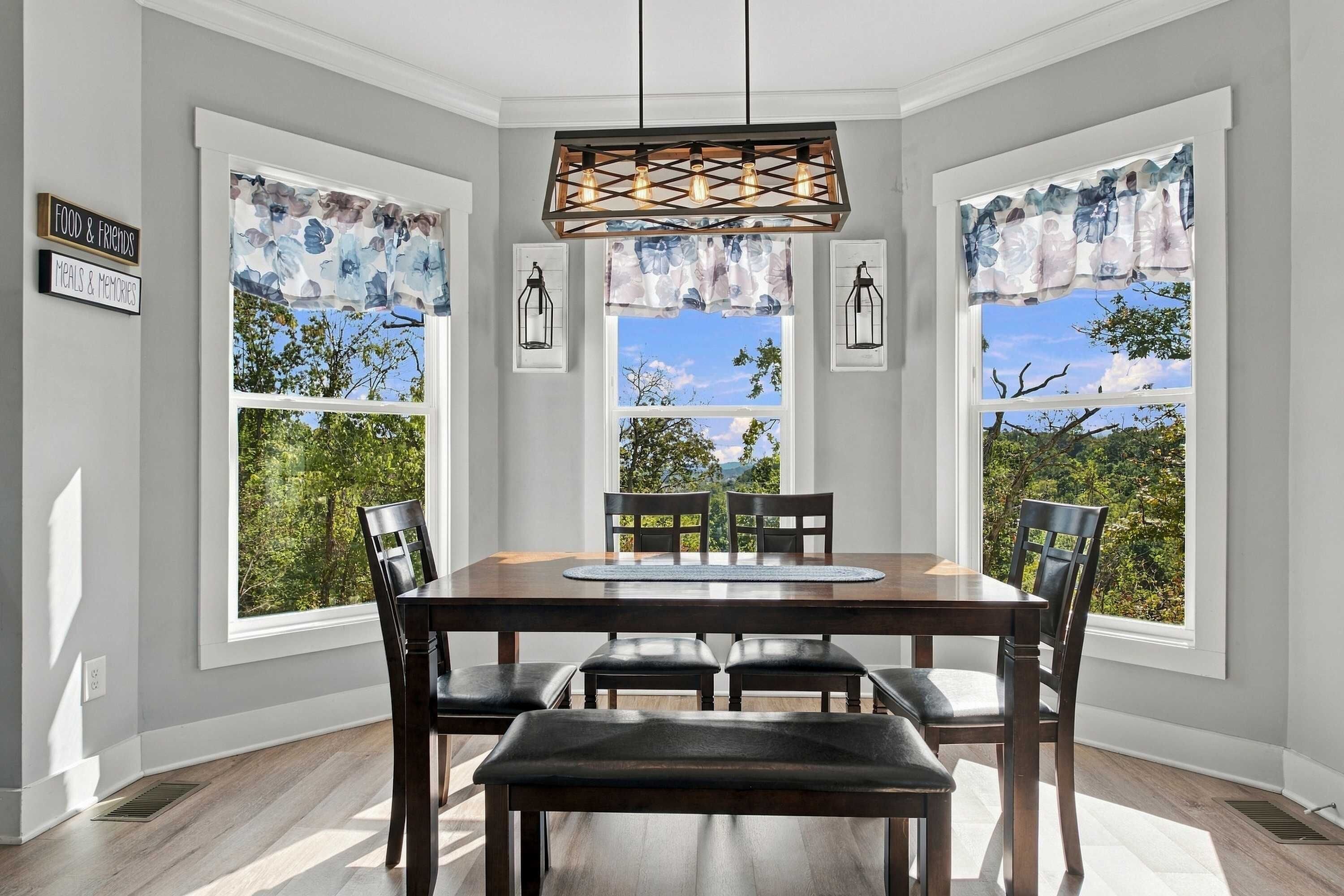 A dining area with a wooden table and chairs, large windows with floral curtains, and a pendant light fixture.
