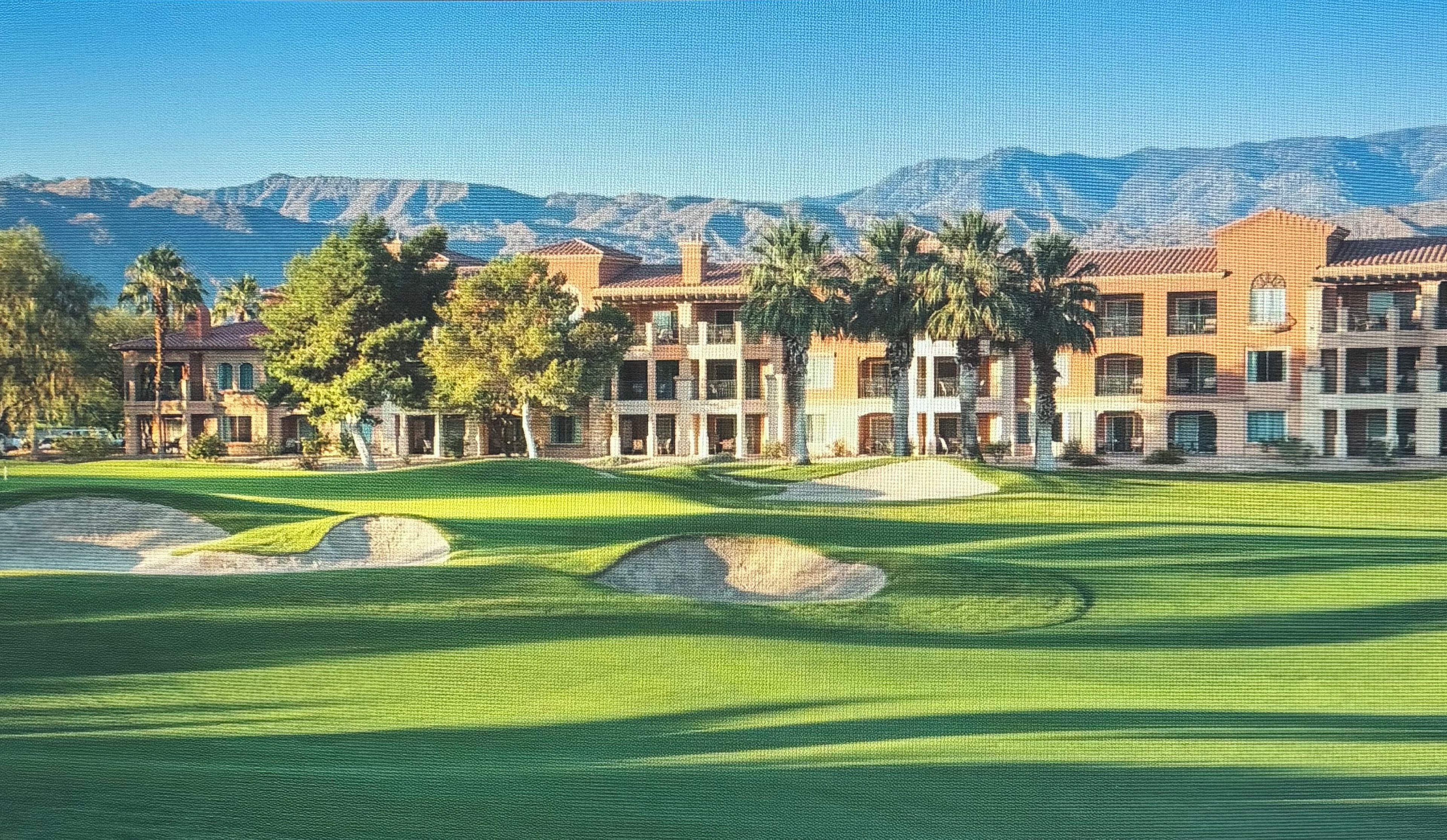 A golf course with multiple buildings in the background, a clear sky, and mountains in the distance.