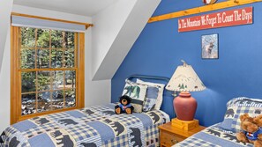 Ein Schlafzimmer mit großem Fenster, einem Bett mit blau-weiß kariertem Überwurf, einem Teddybären, einer Lampe und einem Wandschild.