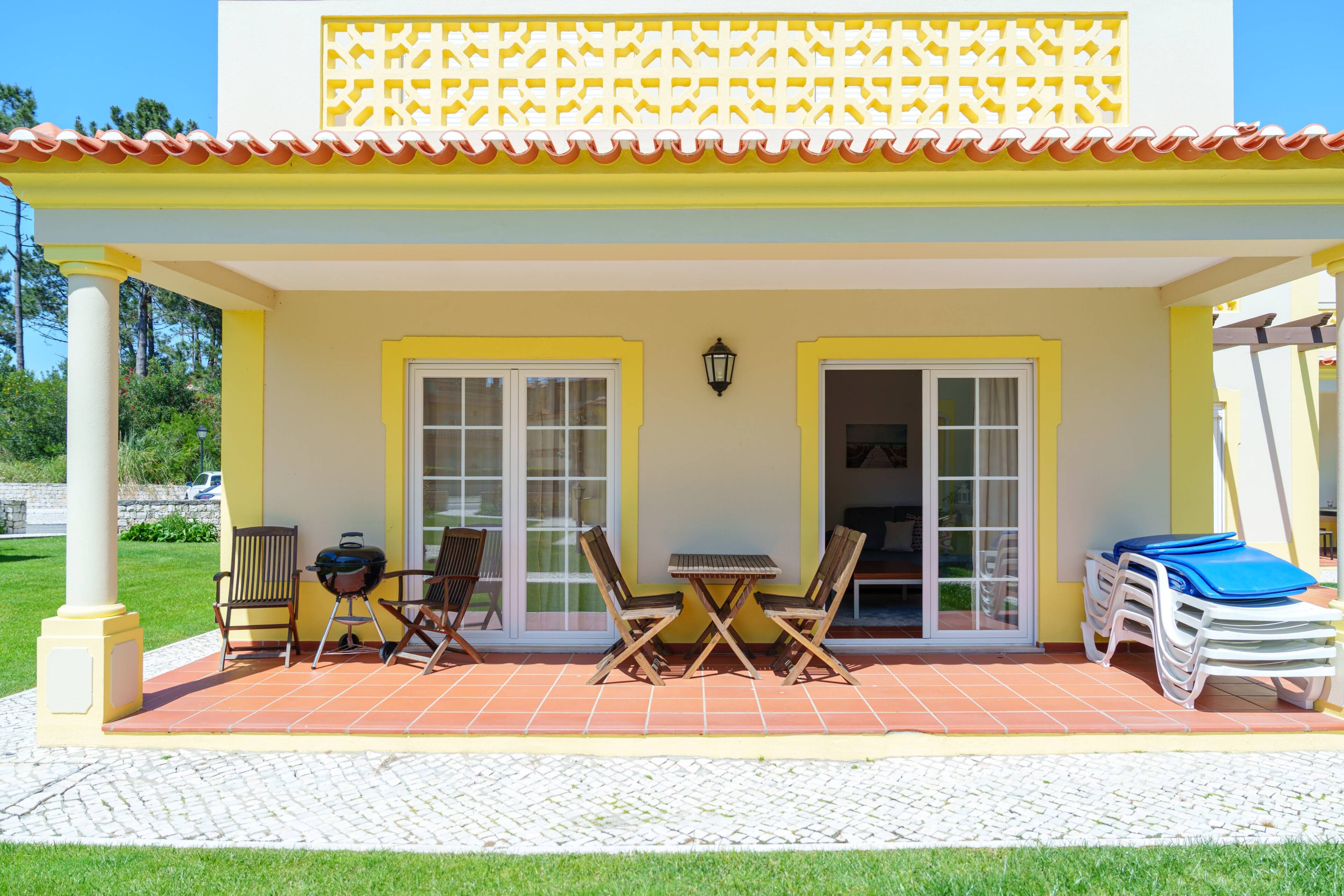 A covered patio area with a table and chairs, a grill, and a stack of lounge chairs.