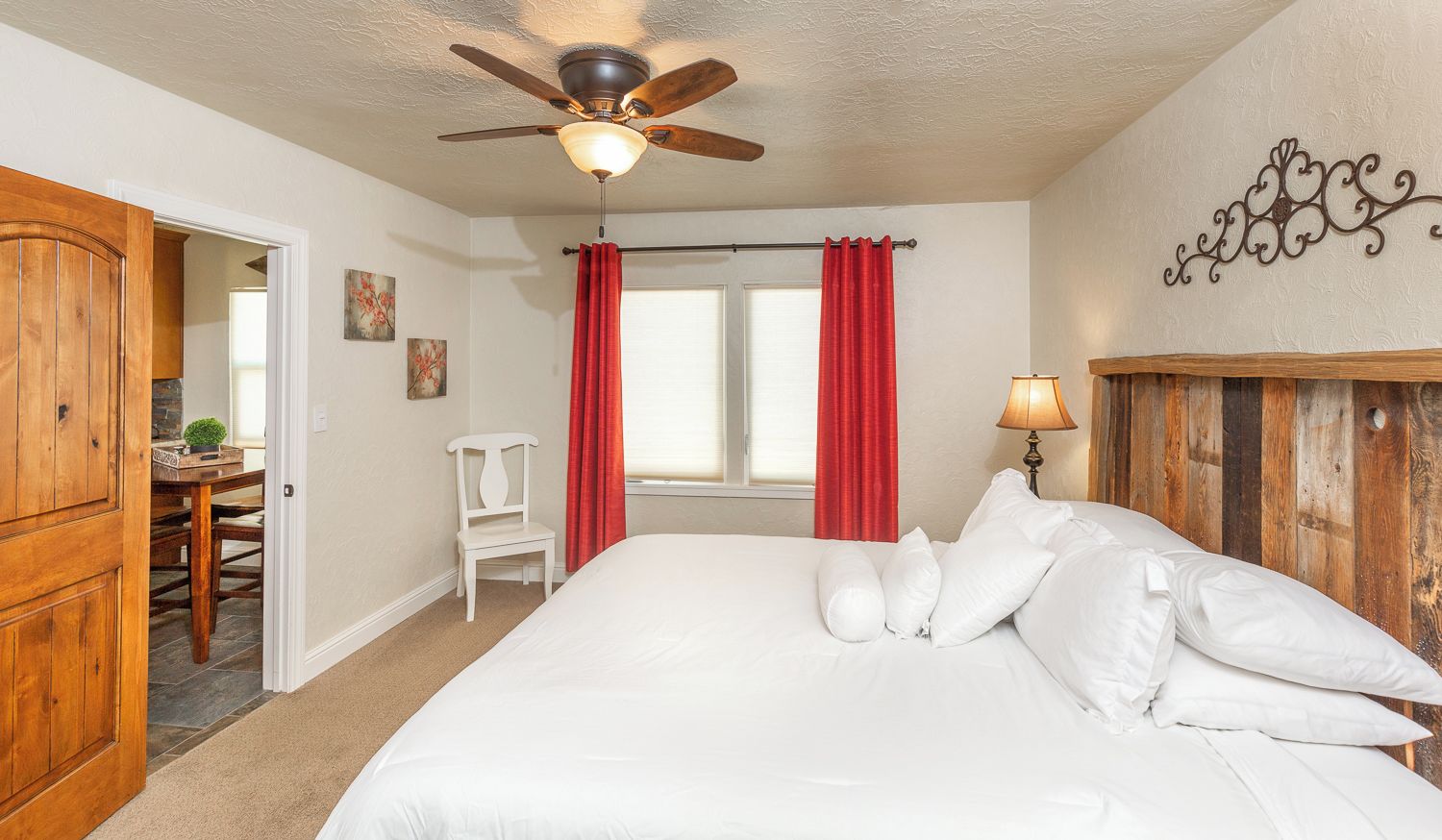 Un dormitorio con cama, ventilador de techo, ventana con cortinas, cabecera de madera y lámpara de pared.