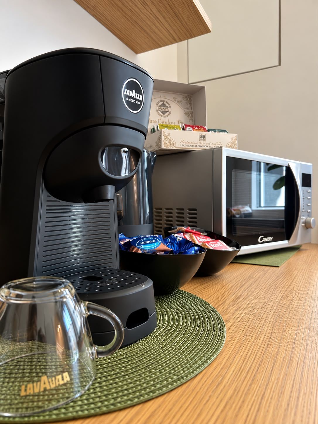 A coffee maker, a microwave, a bowl of snacks, and a glass on a table.