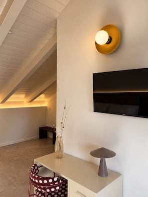 Ein modernes Interieur mit einem weißen Schreibtisch, einer Vase mit Zweigen, einem stippelten Stuhl, einer Wandlampe und einem Flachbildfernseher.
