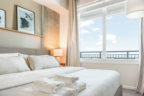 Una camera d'albergo con un letto, un comodino, una lampada, una finestra vista mare e opere d'arte incorniciate al muro.