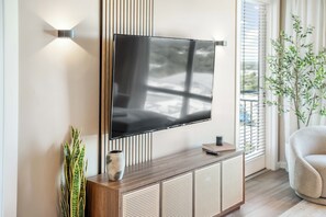 A modern living room with a flat-screen TV mounted on the wall, a wooden console table with a vase and remote control, and a potted plant. - The Louie - Luxe 1BR Condo DT w/ Rooftop Pool, Gym (Orlando)