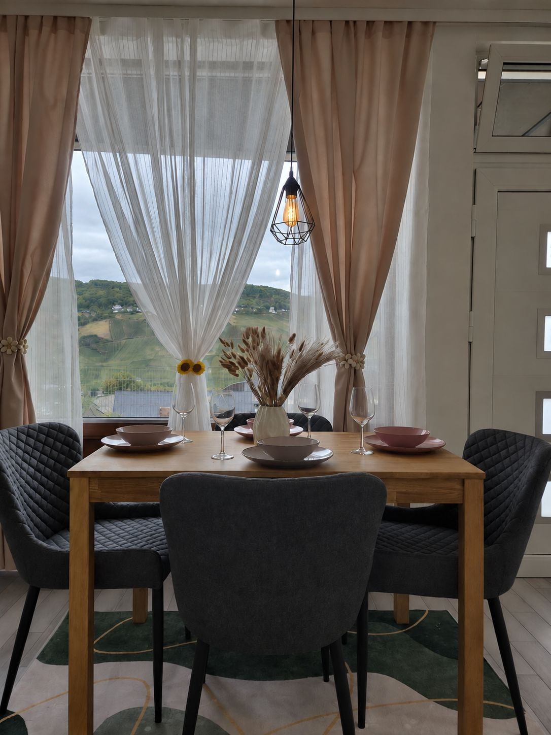 A dining area with a table set for two, a vase with dried flowers, and a view of green hills through sheer curtains.