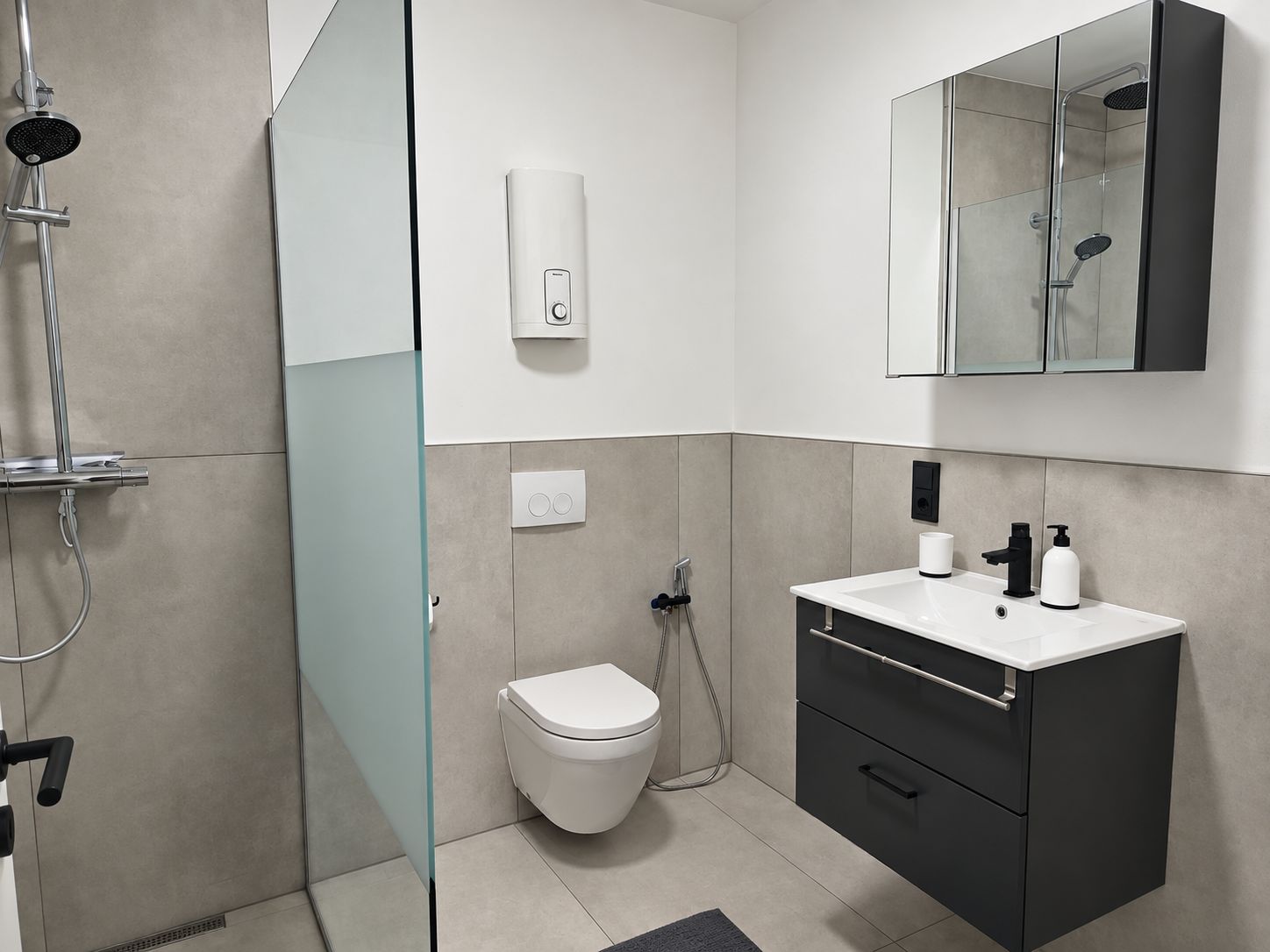 A modern bathroom with a glass shower enclosure, a toilet, a vanity with a mirror, and a wall-mounted soap dispenser.