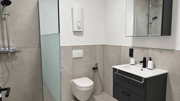 A modern bathroom with a glass shower enclosure, a toilet, a vanity with a mirror, and a wall-mounted soap dispenser.
