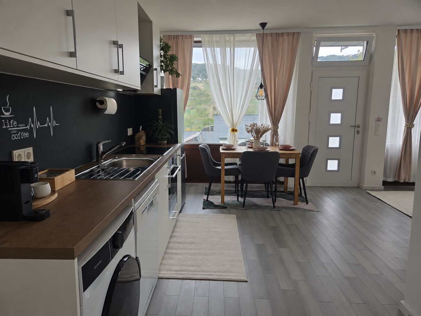 A modern kitchen with a built-in dishwasher, a dining area with a table and chairs, and a view through a window.