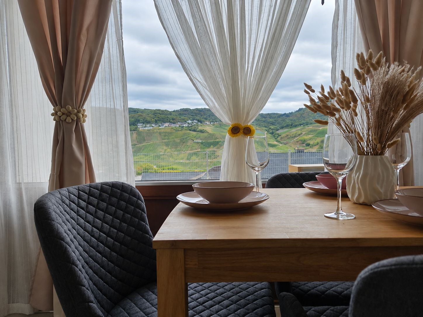 A dining area with a table set for two, a view of the countryside, and a window with sheer curtains.