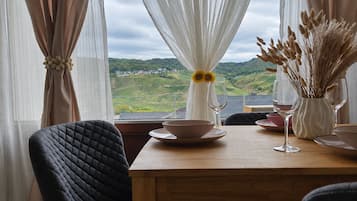 A dining area with a table set for two, a view of the countryside, and a window with sheer curtains.