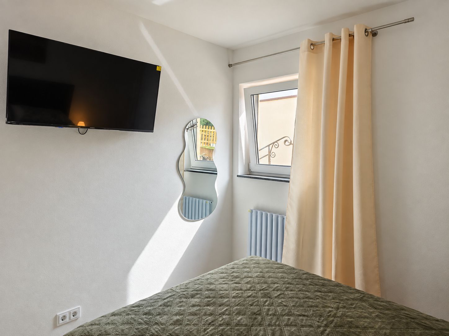 A bedroom with a bed, a mounted TV, a window with curtains, and a radiator.
