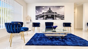 A modern living room with a glass coffee table, blue velvet chairs, a blue shag rug, and a large Eiffel Tower artwork on the wall. - Private Pool BBQ Outdoor Dining (Hollywood)