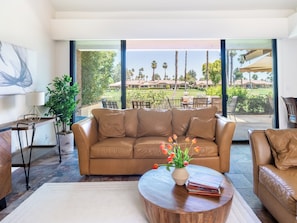 A modern living room with a leather sofa, a wooden coffee table, and a view of a landscaped outdoor area. - Country Club Condo w/ Fairway Views + 37 Pools (Palm Desert)