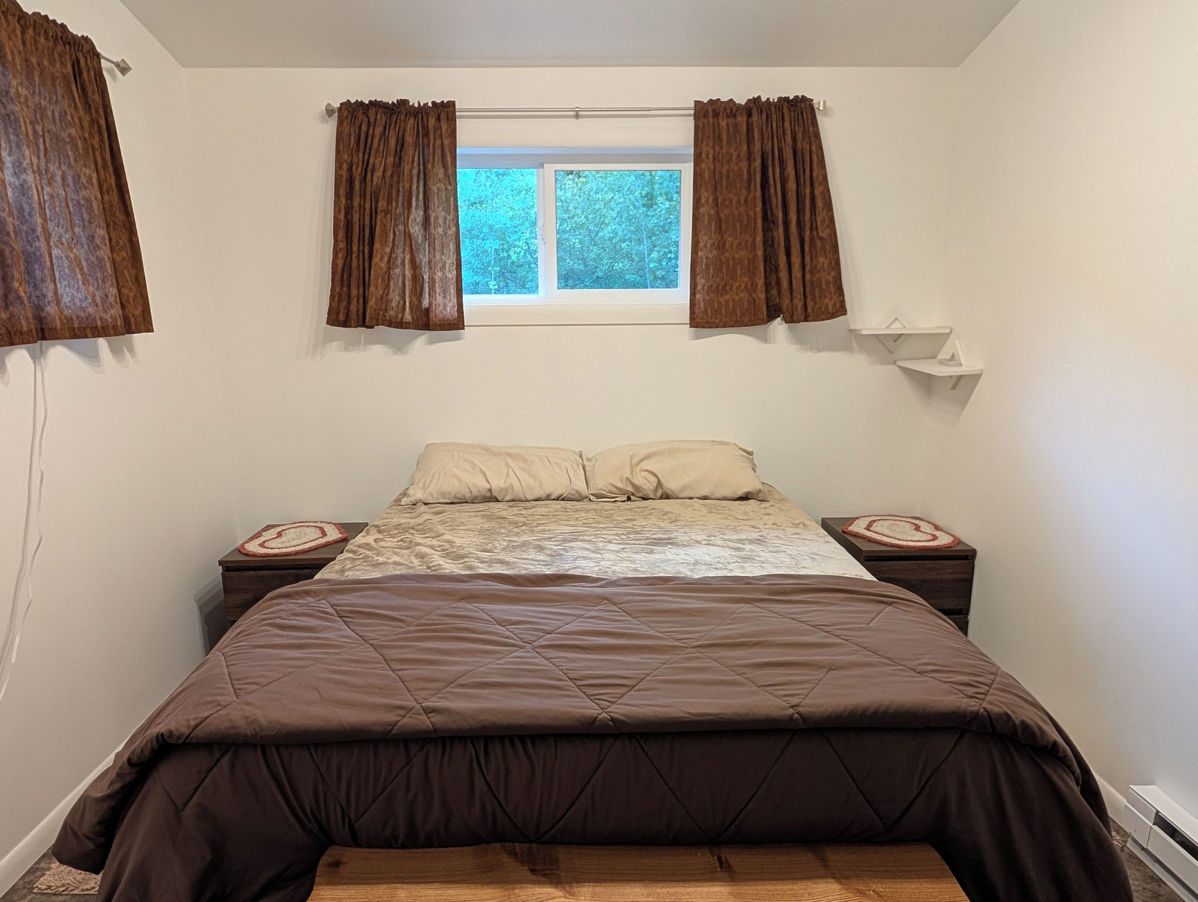A bedroom with a bed, two bedside tables, a window with curtains, and a wall-mounted shelf.