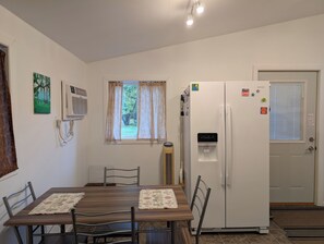 A compact kitchen with a dining table, chairs, a refrigerator, a water dispenser, and a window with curtains.