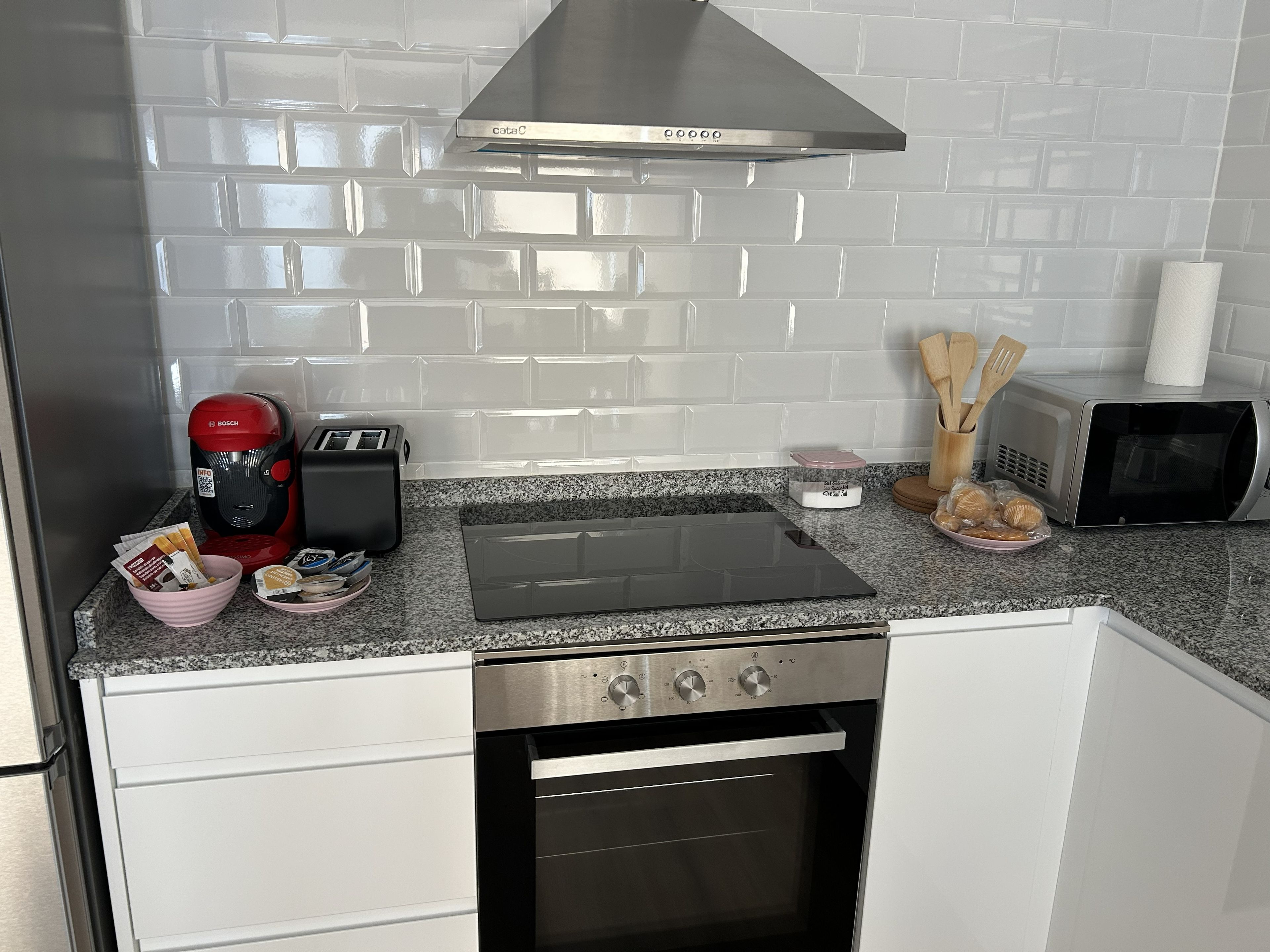 Una cocina con gabinetes blancos, una campana extractora de acero inoxidable, una cafetera Bosch, un tostador, un microondas y una encimera de granito.
