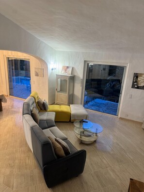 Ein modernes Wohnzimmer mit einem Ecksofa, einem Glastisch und Blick durch eine Glastür auf einen Außenbereich.