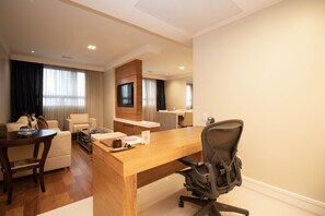 Ein Hotelzimmer mit einem Holztisch, einem schwarzen Bürostuhl, einer Couch, einem an der Wand befestigten Fernseher und einem Essbereich mit Tisch und Stühlen.