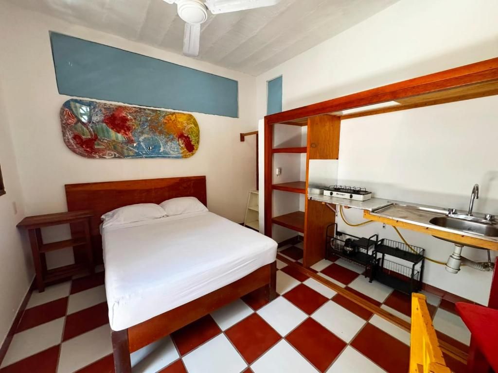 A room with a bed, a small table, a wall-mounted shelf, and a kitchenette area.