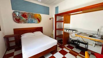 A room with a bed, a small table, a wall-mounted shelf, and a kitchenette area.