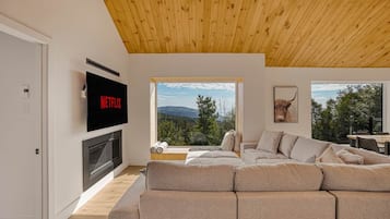 Una sala de estar moderna con un amplio sofá modular, una chimenea, un televisor y ventanas grandes que ofrecen vistas al exterior.